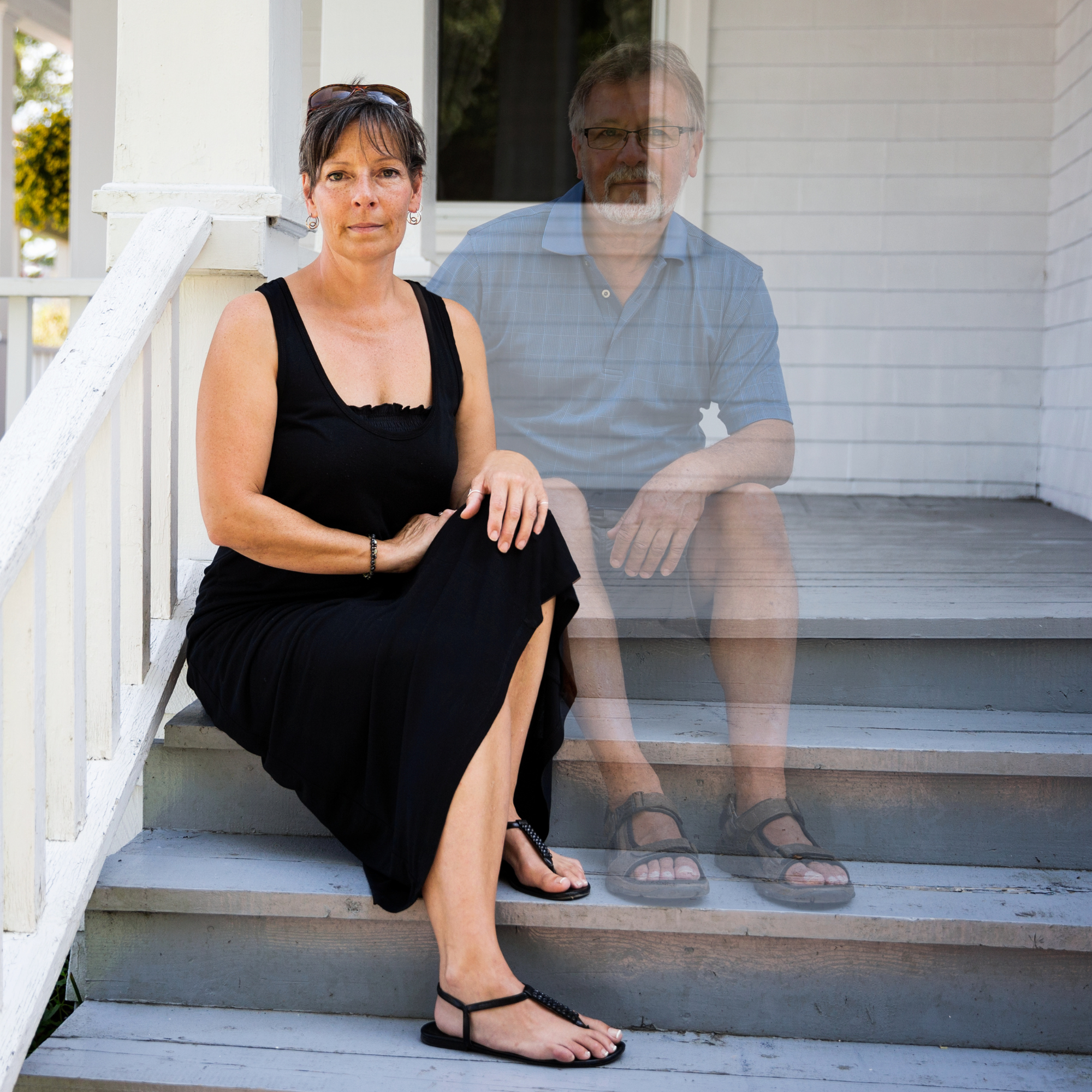 A woman in a black dress, sitting on a porch step, looks directly at the camera. A translucent image of a man sits beside her, suggesting he has recently passed.