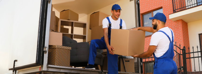 A smiling mover, standing inside a truck, hands a box to another mover.