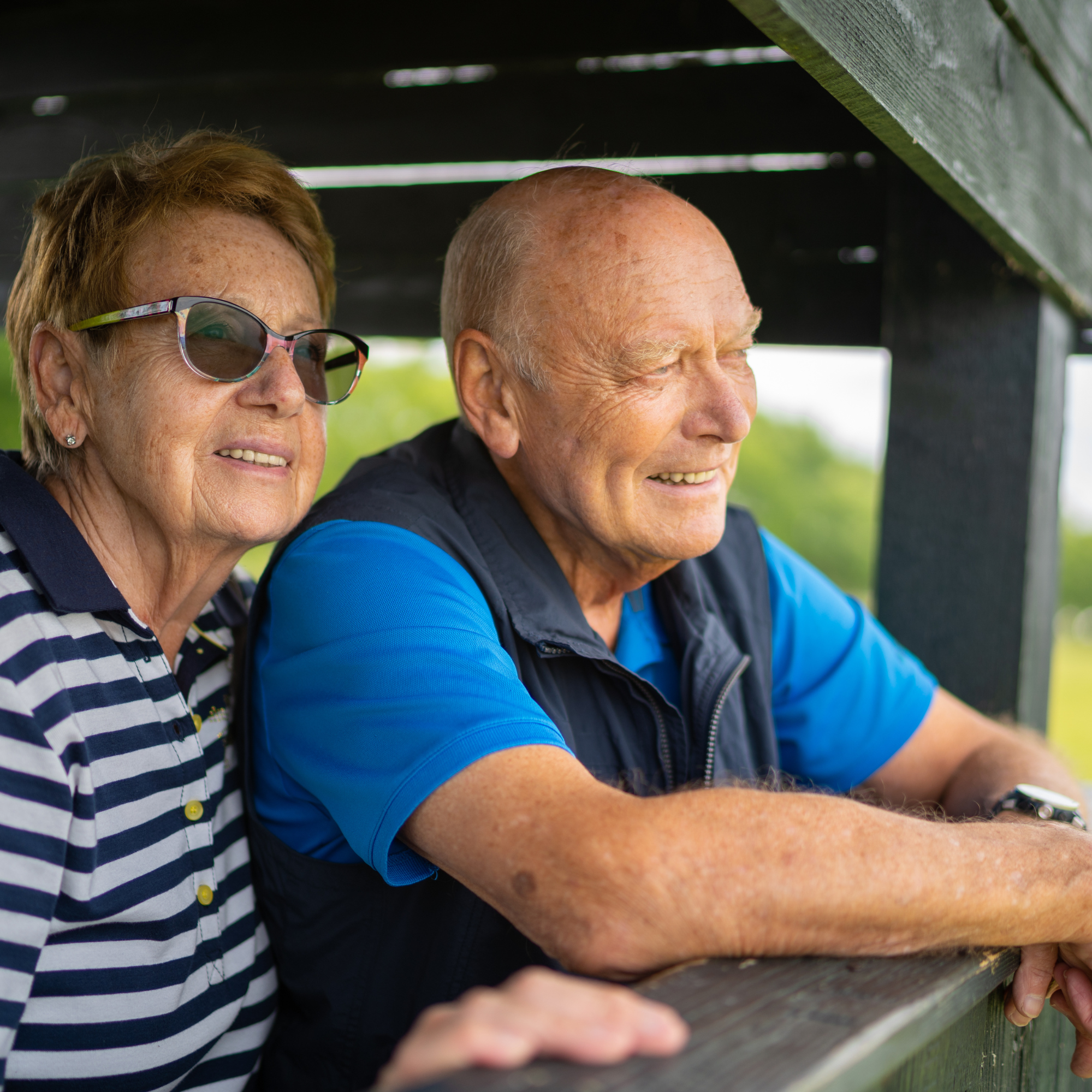 An older couple stands on an outdoor deck or wooden structure, looking off into the distance.