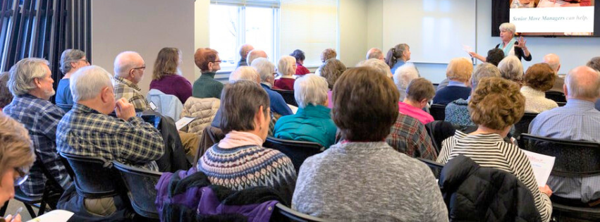 View from the back of an auditorium, full of people sitting in chairs listening to a speaker at the front of the room.
