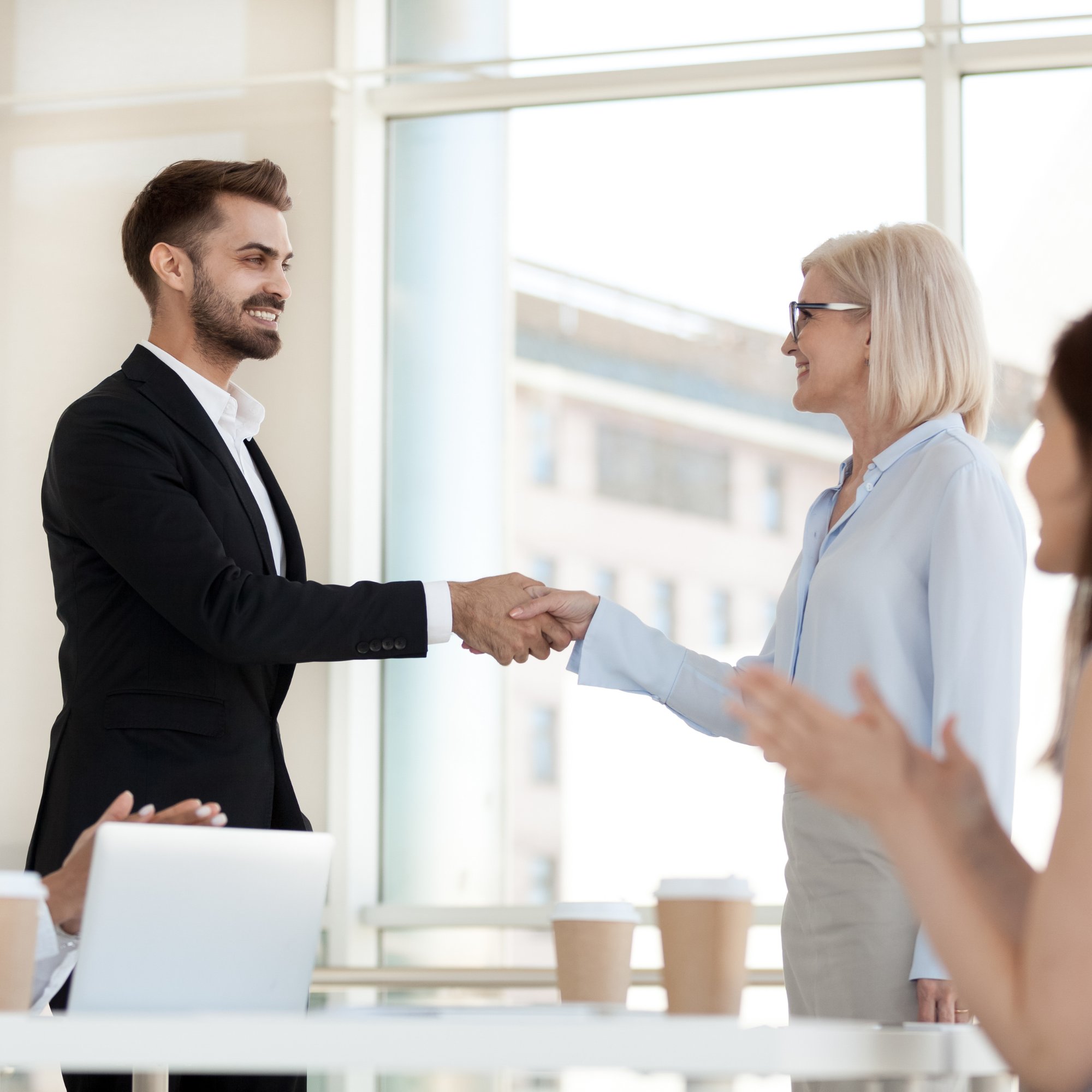 A young man in a black suit shakes hand with a woman in a pale blue blouse and straight blond hair. Clapping hands are visible in the foreground, suggesting perhaps this is an announcement for a promotion.
