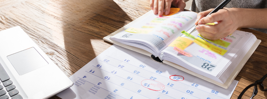 Overhead view of two hands writing in a planner book, with an open calendar and laptop on the table.
