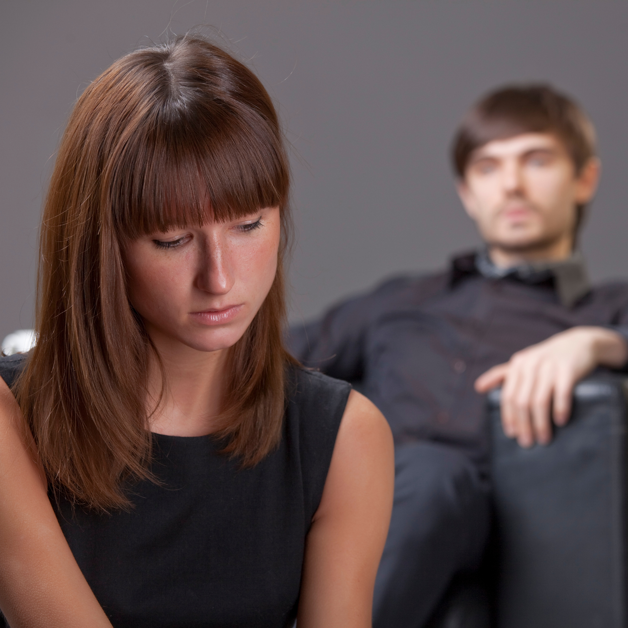 A young woman looks sad in the foreground; a young man sits behind her in a chair, slightly out of focus. It appears they may have just had an argument.